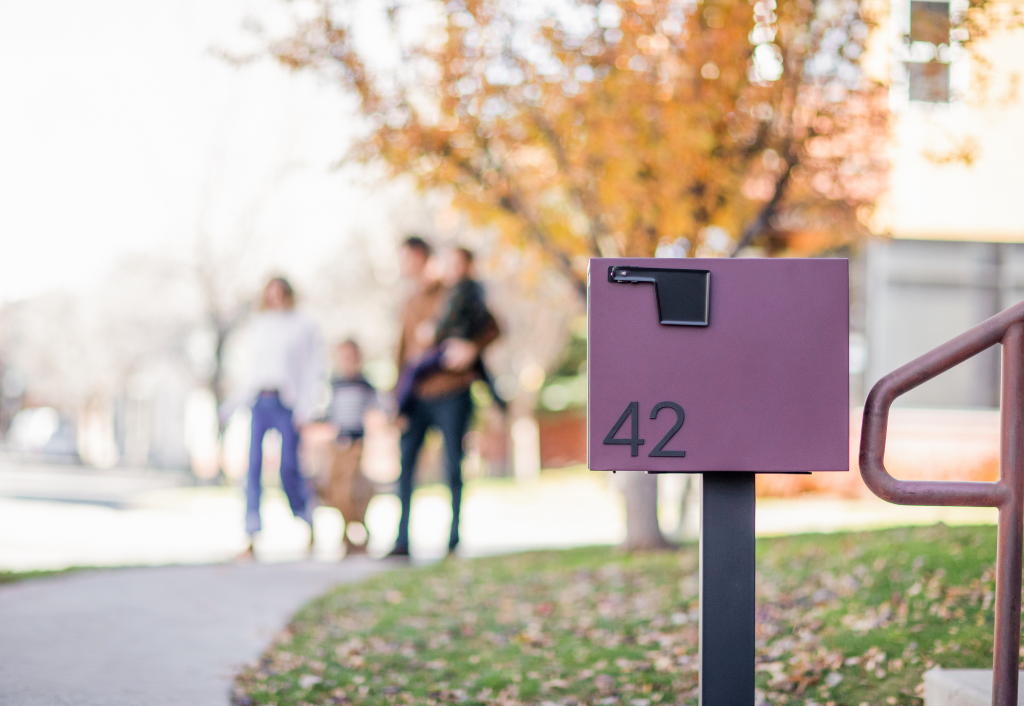 A homeowner at a Malbec Adoorn locking post mount mailbox at the curb, shown in a wide landscape view