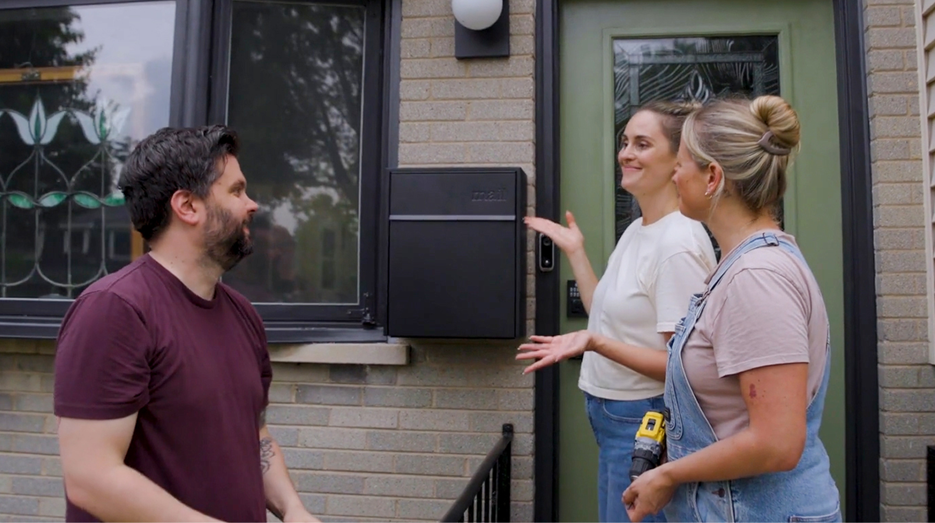 Three people discussing a black Adoorn wall mount mailbox beside a front door