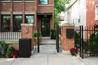 A black Adoorn package box beside a wall mount at a brick home, viewed from the street