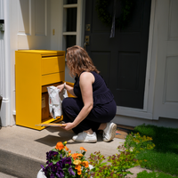 Adoorn Package Box in Curb Yellow on a homeowner's porch, model retrieving a delivery, the always-home alternative to driving to an Amazon Locker.