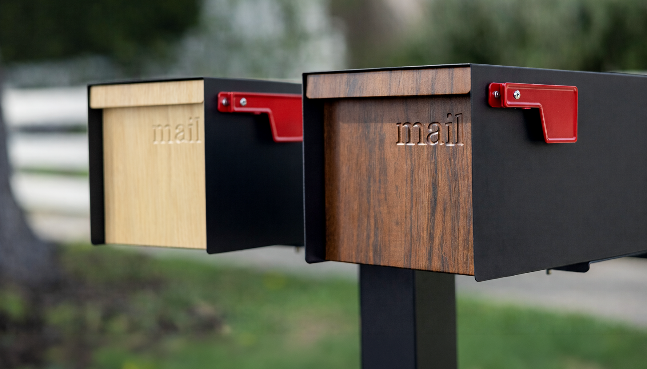 Two Adoorn Wood Series Post Mount mailboxes side by side at the curb, one in white oak and one in walnut, the engraved 'mail' wordmark and red flag visible on each.