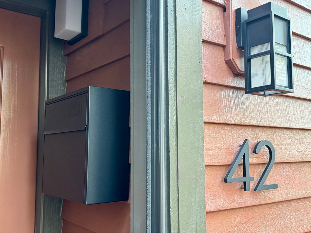 Adoorn black Locking Wall Mount mailbox installed at a covered porch entry with silver 6-inch house numbers
