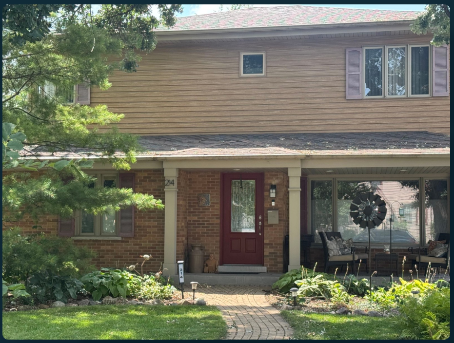 Before: a two-story home with tan siding, red brick, a red front door, porch seating and a curved brick path through garden beds.