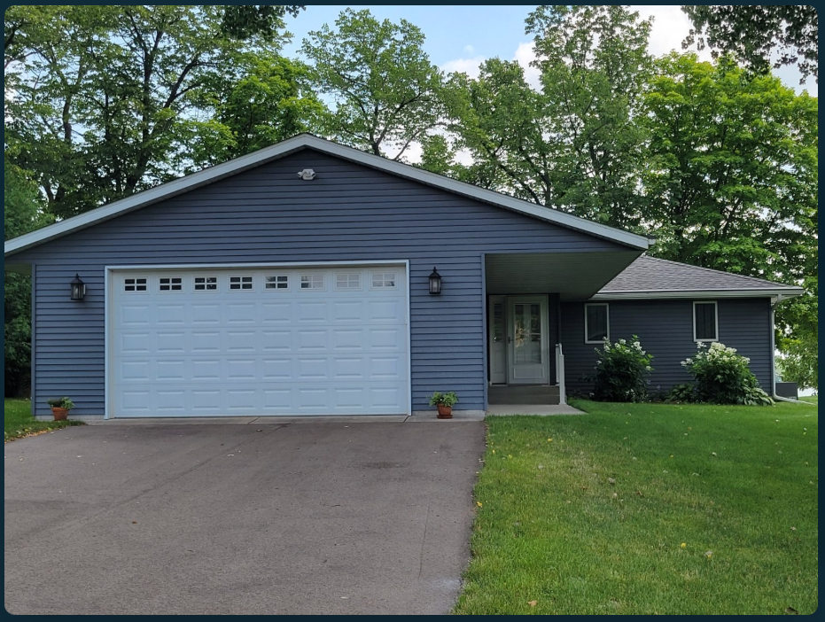 Before: a blue-gray lakeside home with a white garage door, asphalt driveway, recessed entry and lawn bordered by shrubs.